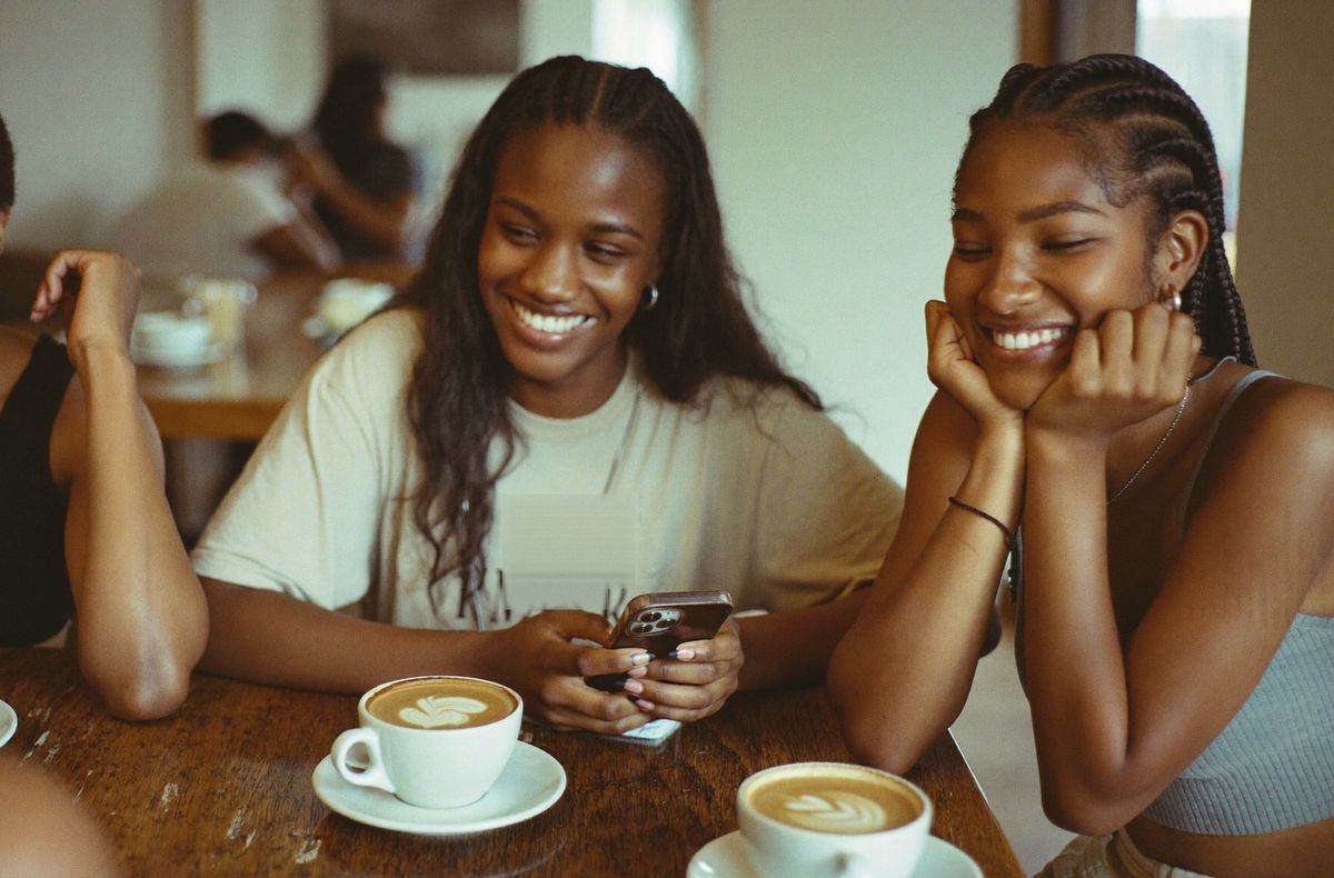 Friends laughing over lattes at a café table, phones out